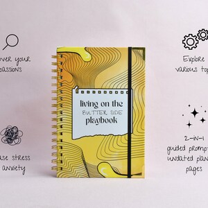 Front view of hard cover of a yellow, guided journal, gold binding, gold corners, elastic holder. Features captions that describe the benefits of the journal: release stress, discover passions, explore various topics, combined journal and planner.