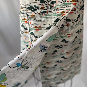 May include: An apron with a landscape pattern of trees, water, and suns in green, brown, and orange. The apron's trim features a white fabric with colorful insect illustrations. The apron is displayed on a mannequin.