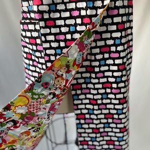 May include: A black and white patterned apron with speech bubbles in pink, blue, and white. The apron's tie features a colorful cartoon design. The apron is displayed on a mannequin.