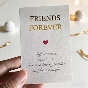 May include: A cream-coloured card with gold lettering that reads "FRIENDS FOREVER." A small red heart is centered below the text. The card also includes the text "Different lives, same heart - here's to late-night talks and forever laughs."