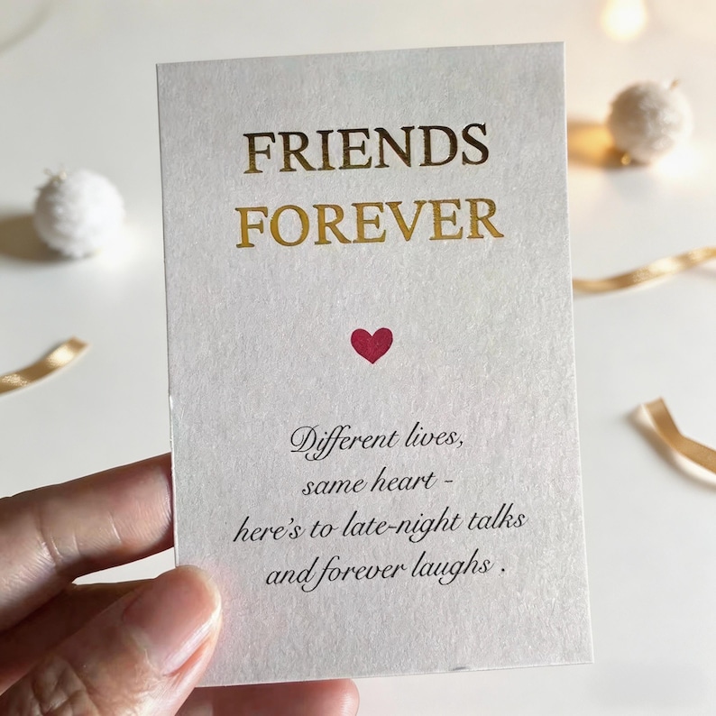 May include: A cream-coloured card with gold lettering that reads "FRIENDS FOREVER." A small red heart is centered below the text. The card also includes the text "Different lives, same heart - here's to late-night talks and forever laughs."