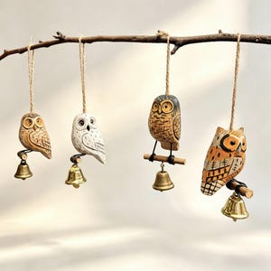 May include: Four carved wooden owl ornaments hanging from a branch. Each owl is a different color and design, with a small brass bell attached below. The owls are suspended by twine.
