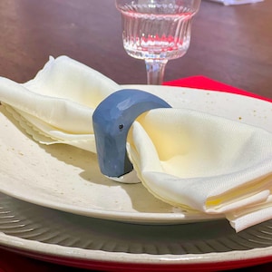 May include: A white napkin is folded and held together by a blue ceramic napkin ring. The napkin ring is shaped like a curved, abstract form. The napkin is on a white plate with a speckled pattern.