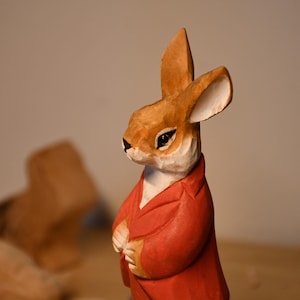 May include: A hand-carved wooden rabbit figurine with a red jacket. The rabbit has brown fur, white accents, and black eyes. The figurine is a detailed representation of a storybook character.