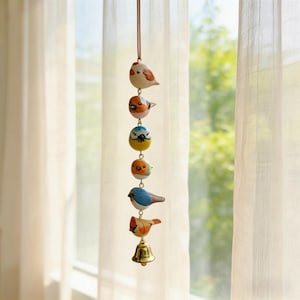 May include: A decorative hanging mobile with six painted bird ornaments in a row. The birds are in shades of red, blue, orange, and yellow, suspended from a brown string. A small gold bell hangs at the bottom of the mobile.