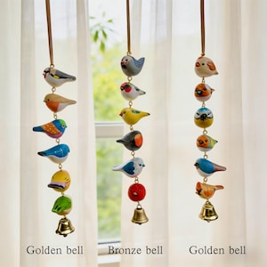 May include: Three hanging bird wind chimes. Each chime features a series of colourful, painted wooden birds strung together, with a bell at the bottom. The chimes are suspended from brown cords. The text "Golden bell" and "Bronze bell" are at the bottom.