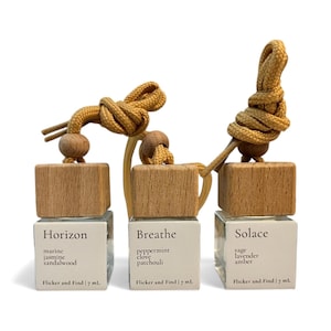 May include: Three glass car air fresheners with wooden tops and tan cords. Each has a white label with a scent name and ingredients: Horizon, Breathe, and Solace. The Horizon scent includes marine, jasmine, and sandalwood.