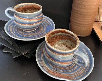May include: Two blue and brown striped ceramic coffee cups with matching saucers. The cups are filled with a light brown substance.