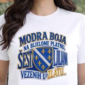 May include: White t-shirt with a blue and gold graphic design. The design includes text and a shield with fleur-de-lis symbols. The text reads "Modra Boja Na Bijelome Platnu, Šest Ljubljana Vezenih U Zlatu."