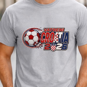 May include: A light gray t-shirt featuring a soccer ball and the text "VATRENI CROATIA 2026" in red, white, and blue. The design includes the Croatian coat of arms and a stylized flame effect.