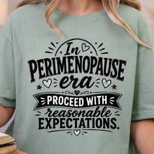 Woman wearing a neutral beige Gildan T-shirt with black In My Perimenopause Era graphic, funny perimenopause and menopause gift for women.