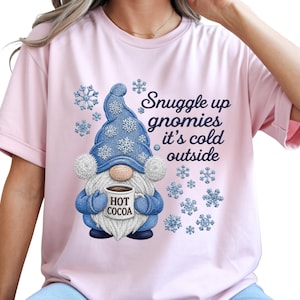 May include: A light pink short-sleeved t-shirt featuring a graphic of a gnome wearing a blue hat with white snowflakes, holding a mug labeled HOT COCOA. The text reads Snuggle up gnomies it's cold outside, surrounded by blue snowflake illustrations.