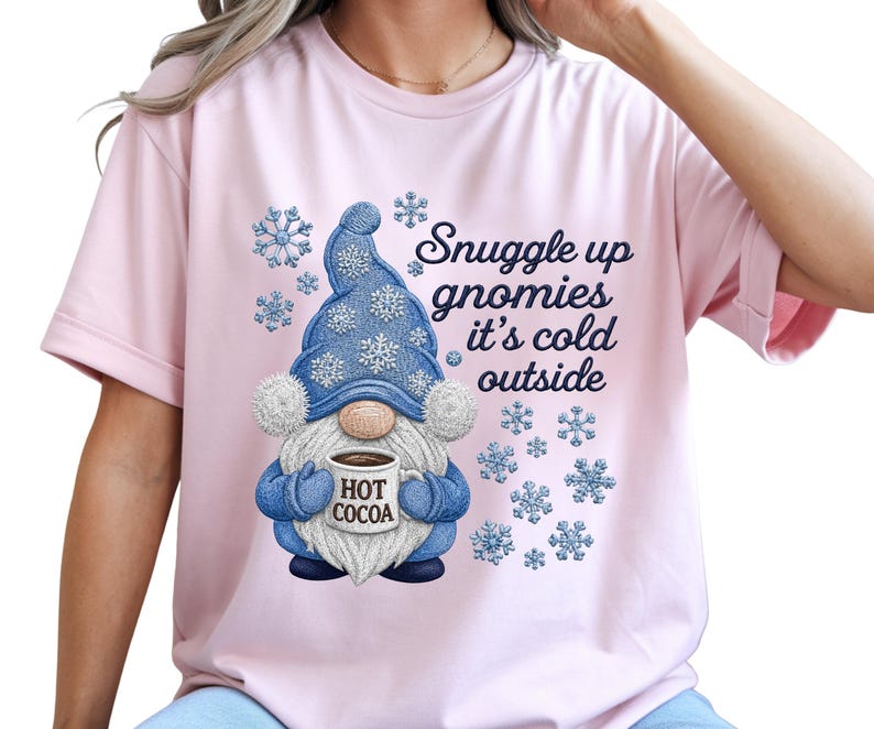 May include: A light pink short-sleeved t-shirt featuring a graphic of a gnome wearing a blue hat with white snowflakes, holding a mug labeled HOT COCOA. The text reads Snuggle up gnomies it's cold outside, surrounded by blue snowflake illustrations.