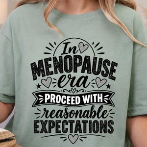 May include: A sage green short-sleeved t-shirt featuring black and silver glitter text that reads In MENOPAUSE era PROCEED WITH reasonable EXPECTATIONS, accented with several small heart shapes and decorative lines throughout the design.