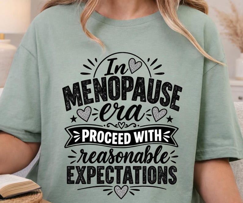 May include: A sage green short-sleeved t-shirt featuring black and silver glitter text that reads In MENOPAUSE era PROCEED WITH reasonable EXPECTATIONS, accented with several small heart shapes and decorative lines throughout the design.