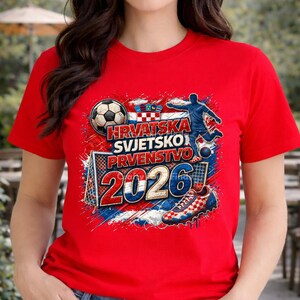 May include: Red t-shirt featuring a soccer-themed graphic. The design includes a soccer ball, a soccer player, a goal, and the text "Hrvatska Svjetsko Prvenstvo 2026" in white and blue. The shirt has short sleeves.