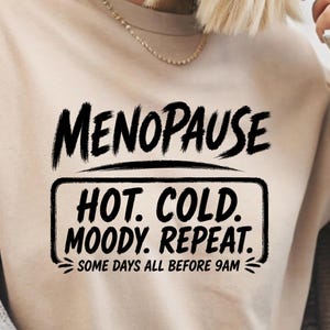 May include: A beige t-shirt featuring black text that reads MENOPAUSE, followed by a rectangular box containing the words HOT. COLD. MOODY. REPEAT. and the additional text SOME DAYS ALL BEFORE 9AM at the bottom in a bold, brush-stroke style font.