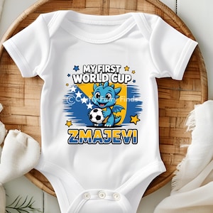 May include: White baby bodysuit with a graphic featuring a blue dragon holding a soccer ball. The design includes the text "MY FIRST WORLD CUP" and "ZMAJEVI" with the colors of the Bosnian flag. The bodysuit is on a woven basket.
