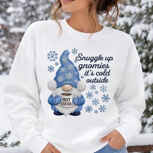 May include: A white crewneck sweatshirt featuring a graphic of a gnome wearing a blue hat with snowflake patterns, holding a mug labeled HOT COCOA. The text reads Snuggle up gnomies it's cold outside, surrounded by blue snowflake illustrations.