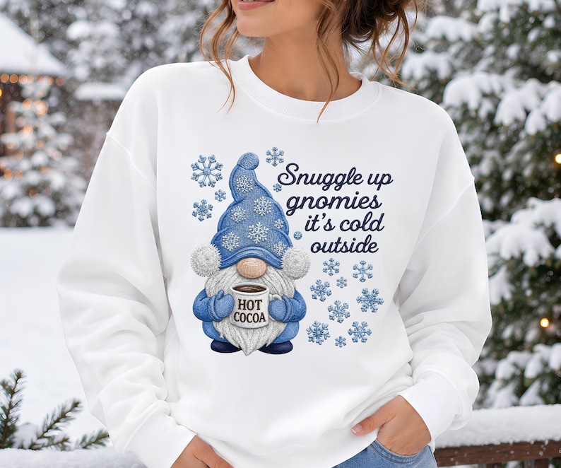 May include: A white crewneck sweatshirt featuring a graphic of a gnome wearing a blue hat with snowflake patterns, holding a mug labeled HOT COCOA. The text reads Snuggle up gnomies it's cold outside, surrounded by blue snowflake illustrations.