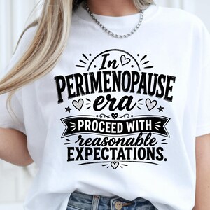 May include: A white short-sleeved t-shirt featuring black text that reads In PERIMENOPAUSE era PROCEED WITH reasonable EXPECTATIONS. The design includes decorative elements such as small hearts, stars, and a banner graphic.