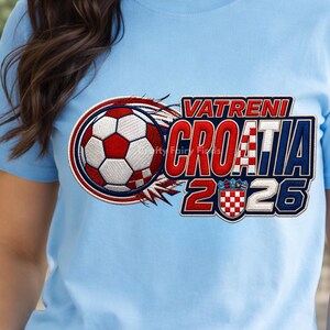 May include: Light blue t-shirt featuring a soccer ball with red and white accents, and the text "VATRENI CROATIA 2026". The design includes the Croatian coat of arms. The shirt is made of a soft material.