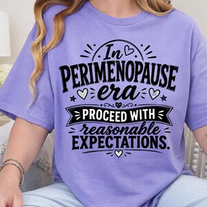 May include: A light purple short-sleeve t-shirt featuring black text that reads In PERIMENOPAUSE era PROCEED WITH reasonable EXPECTATIONS. The design includes decorative elements such as small hearts, stars, and a banner graphic.