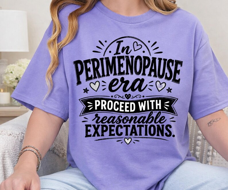 May include: A light purple short-sleeve t-shirt featuring black text that reads In PERIMENOPAUSE era PROCEED WITH reasonable EXPECTATIONS. The design includes decorative elements such as small hearts, stars, and a banner graphic.