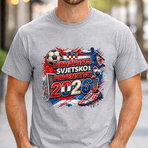 May include: A light gray t-shirt featuring a soccer-themed graphic. The design includes a soccer ball, a soccer player, a goal, and the text "HRVATSKA SVJETSKO PRVENSTVO 2026" in red, white, and blue, representing the Croatian flag.