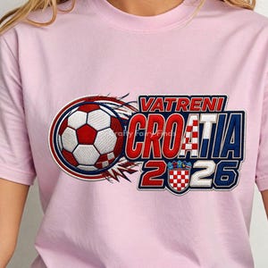 May include: A light pink t-shirt featuring a soccer ball design with red, white, and blue accents. The text "VATRENI CROATIA 2026" is displayed in a bold, stylized font, with a Croatian flag emblem.