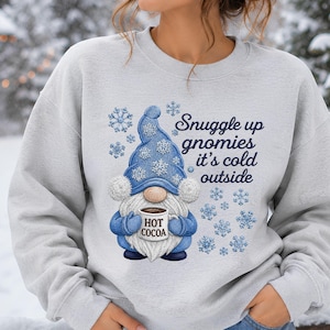 May include: A light gray crewneck sweatshirt featuring a graphic of a gnome wearing a blue hat with white snowflakes. The gnome holds a mug labeled HOT COCOA. The text reads Snuggle up gnomies it's cold outside, surrounded by blue snowflakes.