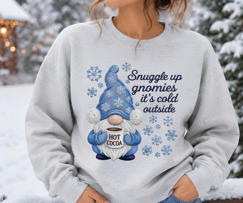 May include: A light gray crewneck sweatshirt featuring a graphic of a gnome wearing a blue hat with white snowflakes. The gnome holds a mug labeled HOT COCOA. The text reads Snuggle up gnomies it's cold outside, surrounded by blue snowflakes.