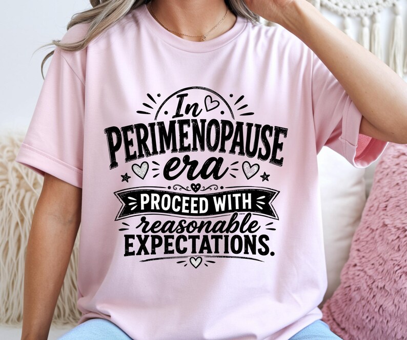 May include: A light pink short-sleeved t-shirt featuring black text that reads In PERIMENOPAUSE era PROCEED WITH reasonable EXPECTATIONS. The design includes small heart and star icons surrounding the lettering in a centered graphic layout.
