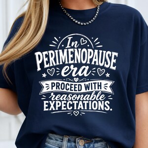 May include: A navy blue short-sleeved t-shirt featuring white graphic text that reads In PERIMENOPAUSE era PROCEED WITH reasonable EXPECTATIONS. The design includes small heart and star accents surrounding the lettering on the front of the shirt.