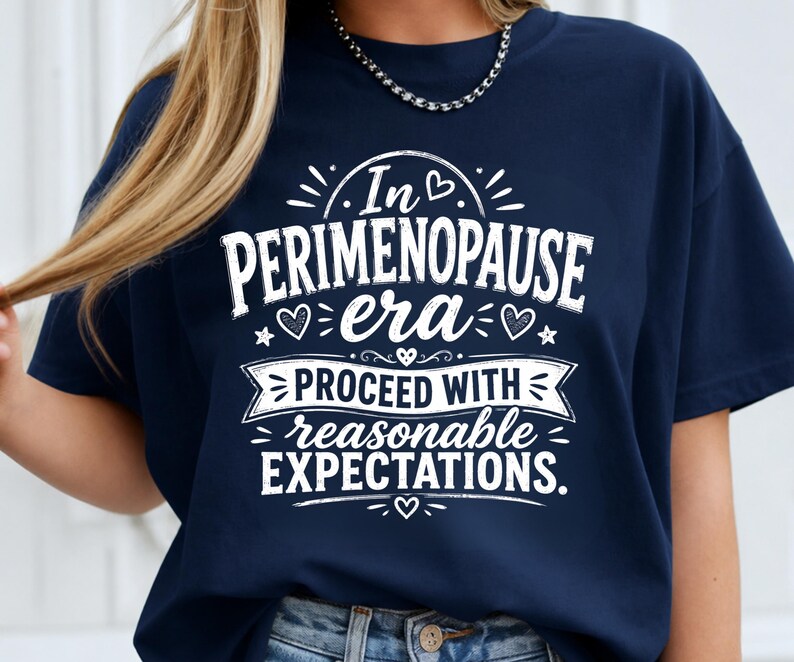 May include: A navy blue short-sleeved t-shirt featuring white graphic text that reads In PERIMENOPAUSE era PROCEED WITH reasonable EXPECTATIONS. The design includes small heart and star accents surrounding the lettering on the front of the shirt.