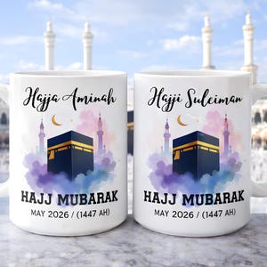 May include: Two white ceramic mugs. One mug has the name "Hajja Aminah" and the other "Hajji Suleiman". Both mugs feature a Kaaba design, a crescent moon, and the text "HAJJ MUBARAK" and "MAY 2026 / (1447 AH)".