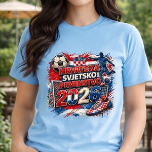 May include: Light blue t-shirt with a graphic design featuring a soccer ball, a soccer player, and a soccer shoe. The design includes the text "HRVATSKA SVJETSKO PRVENSTVO 2026" in red, white, and blue, with the Croatian flag.