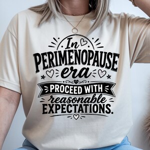 May include: A cream-colored short-sleeve t-shirt featuring black text that reads In PERIMENOPAUSE era PROCEED WITH reasonable EXPECTATIONS. The design includes decorative elements such as small hearts, stars, and lines surrounding the lettering.