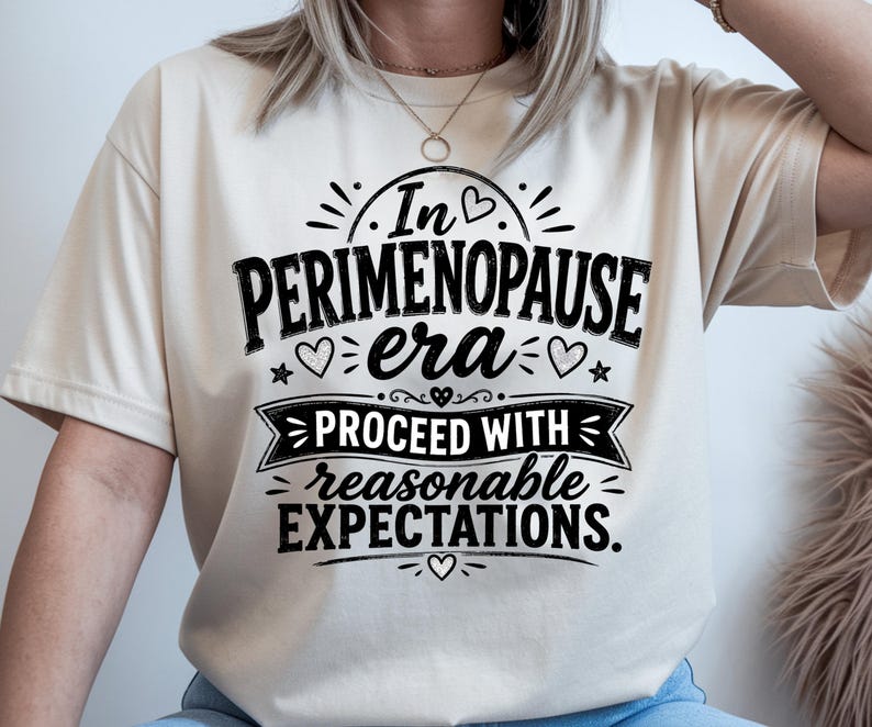 May include: A cream-colored short-sleeve t-shirt featuring black text that reads In PERIMENOPAUSE era PROCEED WITH reasonable EXPECTATIONS. The design includes decorative elements such as small hearts, stars, and lines surrounding the lettering.
