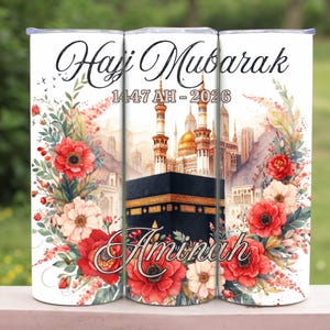 May include: A white travel mug featuring the words "Hajj Mubarak" in script, with the date 1447 AH - 2026. The design includes the Kaaba, a mosque, and the name "Aminah". Red and white flowers and green leaves frame the central image.