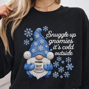 May include: A black long-sleeve sweatshirt featuring a graphic of a gnome wearing a blue hat with white snowflakes. The gnome holds a mug labeled HOT COCOA. The text reads Snuggle up gnomies it's cold outside, surrounded by blue snowflake icons.