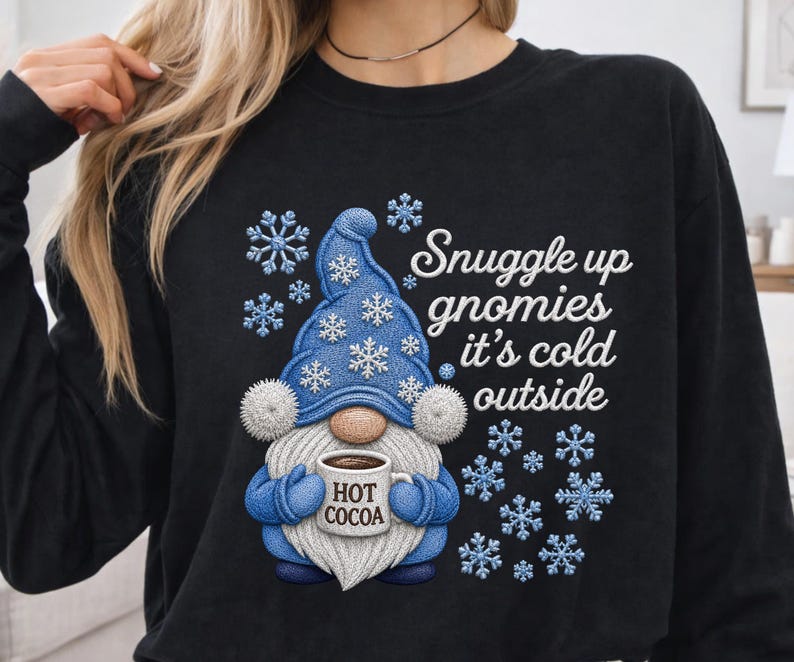 May include: A black long-sleeve sweatshirt featuring a graphic of a gnome wearing a blue hat with white snowflakes. The gnome holds a mug labeled HOT COCOA. The text reads Snuggle up gnomies it's cold outside, surrounded by blue snowflake icons.