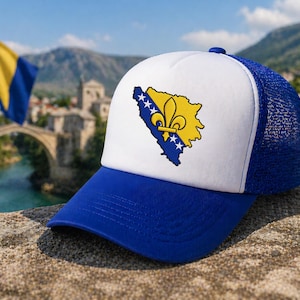May include: A blue and white trucker hat featuring the map of Bosnia and Herzegovina with a yellow fleur-de-lis. The hat has a blue brim and mesh back, with the flag of Bosnia and Herzegovina in the background.