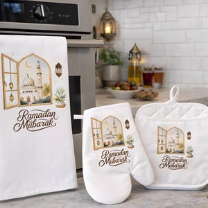 May include: A white kitchen set comprising a tea towel, oven glove, and pot holder. Each item displays a watercolour illustration of a mosque scene within a window, with the words "Ramadan Mubarak". The set is designed for kitchen use.