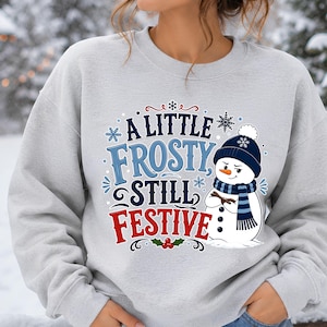 May include: A light gray crewneck sweatshirt featuring a graphic of a snowman wearing a dark blue beanie and scarf. The text reads A LITTLE FROSTY STILL FESTIVE in blue and red lettering, surrounded by small blue snowflakes and red holly berries.