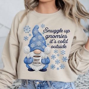 May include: A beige crewneck sweatshirt featuring a graphic of a gnome wearing a blue hat and mittens, holding a mug labeled HOT COCOA. The design includes blue snowflakes and the text Snuggle up gnomies it's cold outside in dark blue script.