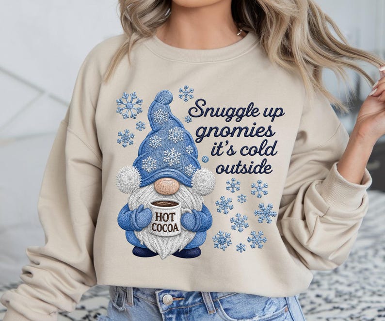 May include: A beige crewneck sweatshirt featuring a graphic of a gnome wearing a blue hat and mittens, holding a mug labeled HOT COCOA. The design includes blue snowflakes and the text Snuggle up gnomies it's cold outside in dark blue script.
