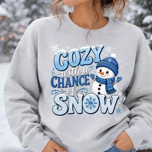 May include: A light gray crewneck sweatshirt featuring a graphic print with the text COZY with a CHANCE of SNOW in blue lettering. The design includes a smiling snowman wearing a blue knit hat and scarf, surrounded by white snowflakes.