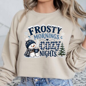 May include: A beige crewneck sweatshirt featuring a graphic print of a snowman wearing a blue plaid hat and scarf, holding a mug. The text reads Frosty Mornings Cozy Nights in blue and white lettering, accompanied by a small green pine tree.