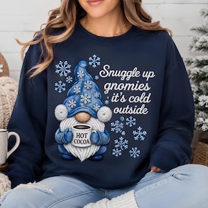 Snuggle Up Gnomies It's Cold Outside shirt and sweatshirt featuring an adorable blue winter gnome holding a hot cocoa mug surrounded by snowflakes. Available as a Gildan 100% cotton long sleeve shirt or 50/50 crewneck sweatshirt in a comfortable unisex true-to-size fit. Adult S–3XL with hoodies, youth, toddler and baby sizes available upon request. Design is a flat DTF print with a simulated embroidered appearance and is not actual embroidery or fabric stitching. Handmade and pressed to order.